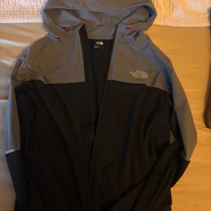 Svart och grå vindjacka/kofta The North Face - vindjacka även som kofta från The North Face i svart och grått. Jackan har huva, dragkedja framtill och logga på bröstet. Perfekt för blåsiga dagar och enkel att matcha med streetwear. Materialet är lätt och känns sportigt.