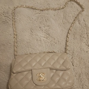 Beige quiltad väska - Supersnygg beige axelväska quiltat skinn med klassiskt CC-spänne i guld. Kedjerem med skinninslag ger en lyxig vibe. Perfekt storlek för det viktigaste och ikonisk design som alltid är trendig.