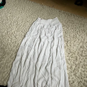 Vit långkjol med resårmidja - Säljer en luftig vit långkjol med bred resår i midjan. Kjolen har en lätt rynkad struktur och är perfekt för varma dagar. Enkel och stilren design som passar till många olika toppar.
