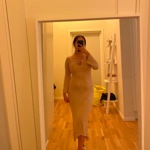 Beige långklänning med ringdetalj - Säljer en beige långklänning som man har ovanför bikini med lång ärm och snygg ringdetalj framtill vid halsen. Klänningen har en kroppsnära passform och är tillverkad i ett mjukt, stretchigt material. Perfekt för dig som gillar stilrena och trendiga plagg.