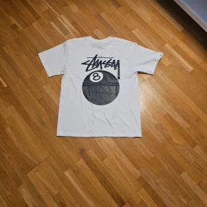 Vit Stüssy t-shirt med 8-ball print - Vit t-shirt från Stüssy i bomull med svart 8-ball print och Stüssy-logga både fram och bak. Klassisk passform med rund hals och korta ärmar. Perfekt för dig som gillar streetwear och vill sticka ut med en ikonisk design.