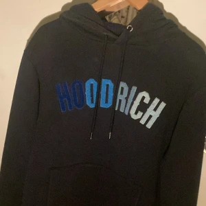 Mörk blå HOODRICH hoodie med blå text - Mörkblå  hoodie från HOODRICH med broderad text i olika blå nyanser över bröstet. Klassisk huva med snörning och stor magficka framtill. Perfekt för dig som gillar streetwear och vill ha en stilren men ändå iögonfallande tröja.