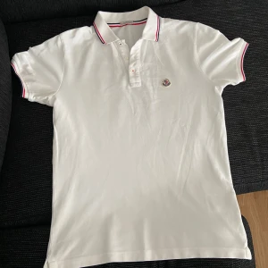 Vit Moncler piké med röd/blå kant - Vit pikétröja från Moncler med klassisk krage och korta ärmar. Tröjan har Moncler-logga på bröstet och snygga röd/blå detaljer på krage och ärmslut. Tillverkad i bomull för skön känsla och stilren look. Perfekt för dig som gillar exklusiva märken.