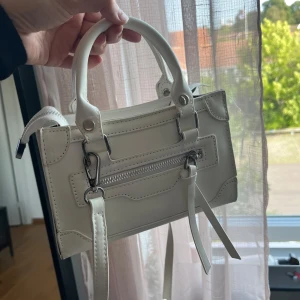 Vit handväska med dragkedja - Säljer en stilren vit handväska i skinnimitation med coola metalldetaljer och dragkedja framtill. Väskan har två handtag och dekorativa remmar som ger en trendig look. Perfekt storlek för det viktigaste och passar till många outfits. Aldrig använd. 