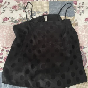 Svart prickig linnetopp från Lindex  - Svart linnetopp från Lindex  med stora prickar i ton-i-ton. Tunt, glansigt material med smala axelband och rak passform. Tillverkad i viskosblandning med LENZING ECOVERO. Perfekt för en stilren och trendig look.