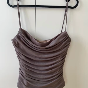 Brun draperad linnetopp från Zara - Säljer en snygg brun linnetopp från Zara med tunna axelband och draperad framsida. Toppen har en croppad passform och är tillverkad i ett mjukt, syntetiskt material som ger en lyxig känsla. Perfekt för dig som gillar stilrena och trendiga plagg.