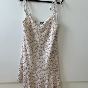 Blommig sommarklänning från Gina Tricot - Supersöt kort klänning från Gina Tricot med smala axelband som knyts upptill. Klänningen är vit med små lila blommor och har en lätt A-linjeformad siluett. Perfekt för varma dagar och ger en romantisk vibe.