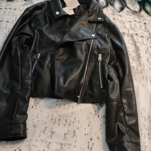 Svart croppad skinnjacka från H&M - Säljer en svart croppad skinnjacka från H&M med coola silvriga dragkedjor och klassisk krage. Jackan är gjord i vattenbaserad PU och har en snygg bikerstil. Perfekt för dig som vill ha en edgy look till din outfit.  Är inte använd pris kan diskuteras
