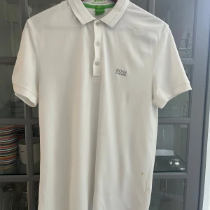 Vit slim fit piké från Hugo Boss - Vit pikétröja från Hugo Boss i slim fit-modell. Klassisk krage med knappar och diskret broderad logga på bröstet. Tillverkad i bomull med fuktavledande egenskaper. Perfekt för en clean och stilren look.