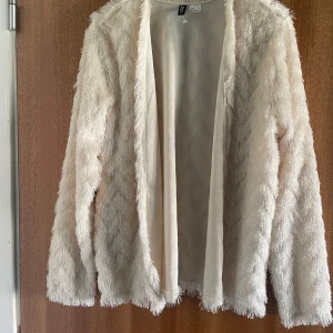 Vit pälsjacka i syntet från H&M - Fluffig vit pälsjacka från H&M i syntetmaterial. Jackan har lång ärm, öppen framtill och en riktigt mjuk, lurvig känsla. Perfekt för dig som vill sticka ut med en cozy look. Passar dig som gillar oversized vibe.