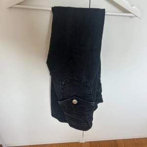 Svarta bootcut jeans med fickdetaljer - Snygga svarta jeans med bootcut passform och coola fickor med lock och silverknappar bak. Jeansen har klassisk femficksdesign och är  tillverkade i ett mjukt denimtyg som sitter skönt. Dom är från Gina tricot och är low waist i storlek 34