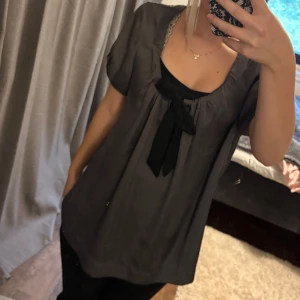 Grå blus med svart rosett från H&M - Säljer en lös och flowy grå blus från H&M med korta ärmar och en snygg svart rosett framtill. Säljer pågrund av att den är för stor😕ser ut att vara helt ny men är osäker eftersom jag köpt den på vinted.