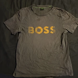 Mörk grå BOSS t-shirt med guld logga - Svart t-shirt från BOSS i storlek XS passar som S med korta ärmar och rund hals. Framsidan har en stor guldig BOSS-logga med coolt randigt mönster. Materialet är mjukt och känns som bomull, perfekt för en chill och stilren look.