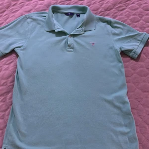 Mintgrön pikétröja från GANT - Fräsch mintgrön pikétröja från GANT med klassisk krage och knäppning framtill. Tröjan har korta ärmar och GANT:s broderade logga på bröstet. Perfekt för en stilren och avslappnad look under vår och sommar.