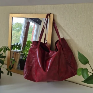 Vinröd axelväska i skinnimitation - Rymlig vinröd axelväska i skinnimitation med långa smala axelband. Väskan har en mjuk, slouchy form och minimalistisk design utan synliga loggor eller detaljer. Supersnygg höstväska🍂Mindre skavank på en rem, visas på bild