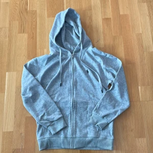 Grå hoodie från Polo Ralph Lauren M - Grå hoodie från Polo Ralph Lauren med dragkedja och huva. Klassisk design med snörning i huvan och broderad logga på bröstet. Tillverkad i mjuk bomullsmix, perfekt för chill dagar eller när du vill ha en clean look.