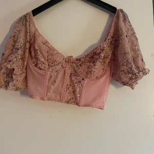 Rosa blommig korsettopp med puffärm - Superfin rosa korsettopp med blommigt mönster och broderade detaljer i guld. Toppen har puffiga, genomskinliga ärmar och en liten rosett framtill. Croppad modell med offshoulder-skärning, perfekt för en söt och romantisk look. (Använd en gång till en halloween kostym) 