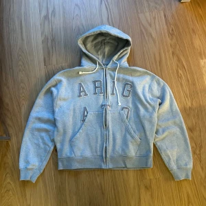 Grå hoodie från Axel Arigato - Säljer en grå hoodie från Axel Arigato med dragkedja och stor broderad logga framtill. Extremt bra kvalité och riktigt tjockt tyg. Köpt på arigato i london, tror jag har kvittot kvar, har kvar alla tags, nypris är £180, digitalt kvitto finns