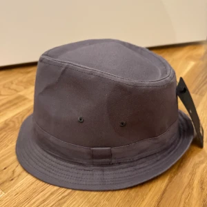Grå hatt från New Yorker - Stilren grå bucket hatt från New Yorker  i bomull. Klassisk form med rundad kulle och nedvikt brätte. Perfekt för att lyfta din streetwear-look eller skydda mot solen. Enkel och clean design utan mönster eller extra detaljer.