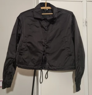 Svart vindjacka med snörning - Svart vindjacka från Weekday med klassisk krage, knappar framtill och snörning i nederkant. Jackan har två sidofickor och är croppad i modellen. Perfekt för dig som gillar stilrena och funktionella plagg.