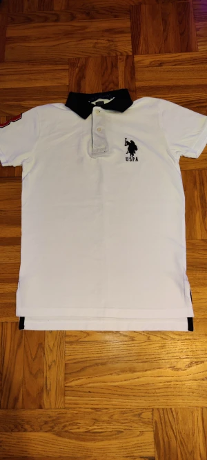 Vit US Polo Assn pikétröja slim fit - Vit pikétröja från US Polo Assn med svart krage och broderad logga på bröstet. Slim fit-modell i 100% bomull. Kortärmad med röd broderad siffra 3 på ärmen och knappar vid halsen. Perfekt för dig som gillar sportig och clean stil.