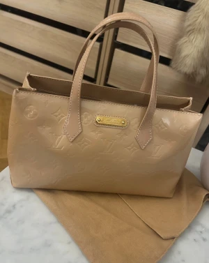 Beige handväska Louis Vuitton Vernis - Snygg beige handväska från Louis Vuitton i Monogram Vernis med glansig finish och präglat LV-mönster. Väskan har två handtag, guldfärgade metalldetaljer och är tillverkad i lackat kalvskinn med textilfoder. Perfekt storlek för det viktigaste och en lyxig touch till din look.