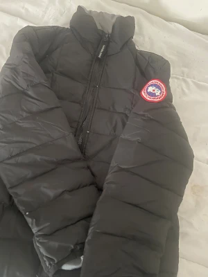 Svart pufferjacka från Canada Goose - Svart pufferjacka från Canada Goose med klassisk logopatch på ärmen. Jackan har hög krage, raka quiltade paneler och hel dragkedja framtill. Perfekt för kalla dagar och har en clean, stilren look som funkar till streetwear.