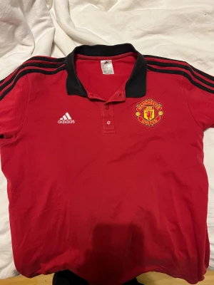 Manchester United röd Adidas fotbollströja - Röd Manchester United fotbollströja från Adidas med svart krage och svarta ränder på axlarna. Broderad klubbmärke på bröstet och Adidas-logga. Klassisk kortärmad modell med knappslå och sportig look. Perfekt för dig som vill representera United.