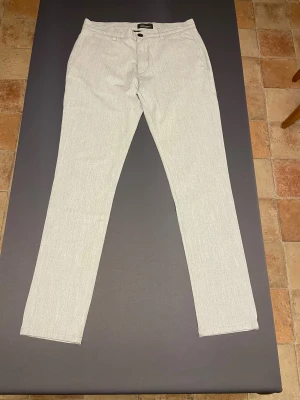 Ljusgrå kostymbyxor Tailored Originals - Snygga ljusgrå kostymbyxor från Tailored Originals med klassisk rak passform och diskreta fickor både fram och bak. Byxorna har bälteshällor, knappstängning och är tillverkade i en mjuk bomullsblandning med polyester och linne för extra komfort.