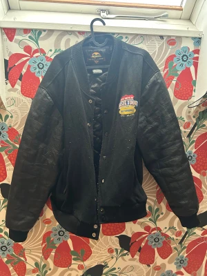 Svart bomberjacka från New Zlander - Säljer en svart bomberjacka från New Zlander med broderad patch på bröstet. Jackan har skinnärmar, ribbad krage och muddar samt tryckknappar framtill. Insidan är quiltad för extra komfort. Perfekt för dig som gillar retro college-vibe. Är köpt på Plick för några år sedan så var redan second hand när jag köpte.