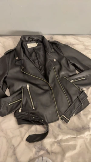 Svart skinnjacka från NLY Trend - Snygg svart skinnjacka från NLY Trend med klassisk bikerstil. Jackan har flera silvriga dragkedjor, knappar och ett bälte i midjan. Modellen är kortare och har krage med tryckknappar. Perfekt för dig som gillar en edgy look.