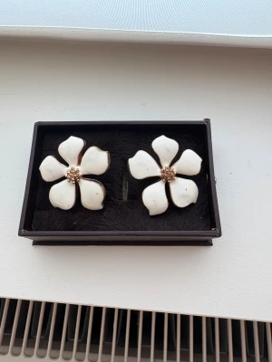 Stora vita blomformade örhängen - Säljer ett par statement örhängen i form av stora blommor med fem kronblad. Framsidan är vit med gulddetaljer i mitten och längs kanterna, baksidan är helguld. Perfekt för dig som vill sticka ut med en cool och unik accessoar.