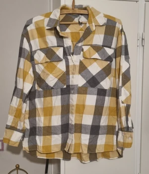 Rutig gul och grå skjorta - Säljer en rutig skjorta i gul, grå och vit med stora rutor. Skjortan har två bröstfickor med lock och knappar framtill. Långärmad och tillverkad i mjuk bomull, perfekt för en chill och avslappnad stil.