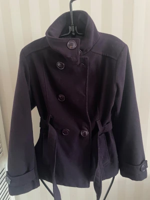 Mörklila skepparkavaj med bälte - Snygg mörklila skepparkavaj med dubbelknäppning och bälte i midjan. Jackan har klassisk krage, långa ärmar med knappdetalj vid mudden och stora fickor framtill. Materialet är mjukt och känns som ull, perfekt för kyliga dagar.