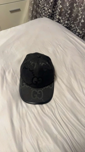 Svart Gucci keps med GG-logga den e äkta  - Säljer en svart keps från Gucci med GG-logga i mörk ton över hela kepsen. Klassisk böjd skärm och justerbar passform. Materialet är polyester och kepsen kommer med originalförpackning och dustbag. Perfekt för dig som vill ha en lyxig streetstyle-accessoar.