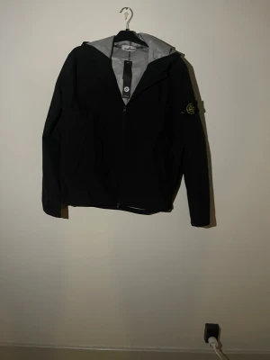 Svart vindjacka från Stone Island - Svart vindjacka från Stone Island med ikonisk patch på vänster ärm. Jackan har huva, hel dragkedja framtill och grått innerfoder. Materialet är syntet och jackan har en clean, normal passform med långa ärmar. Perfekt för dig som gillar streetwear och stilrena detaljer. Storlek M men passar som L.