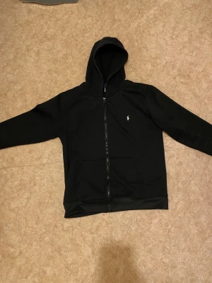 Svart hoodie från Polo Ralph Lauren - Svart hoodie från Polo Ralph Lauren med dragkedja framtill och klassisk logga på bröstet. Tröjan har huva, två fickor och långa ärmar. Perfekt för chill dagar och enkel att matcha med jeans eller joggers.