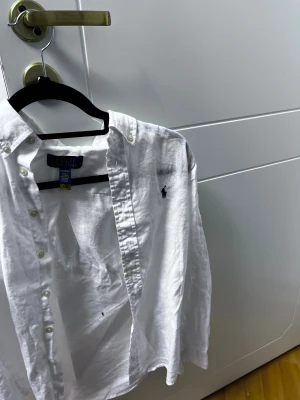 Vit linneskjorta från Polo Ralph Lauren - Fräsch vit skjorta från Polo Ralph Lauren i luftig linne. Klassisk button-down med pärlemorknappar och mörkblå broderad logga på bröstet. Perfekt för dig som gillar clean och stilren look. Skjortan har lång ärm och normal passform.