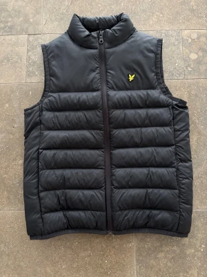 Svart dunväst från Lyle & Scott - Svart dunväst från Lyle & Scott med gul broderad logga på bröstet. Västen har quiltad design, hög krage och dragkedja framtill. Perfekt för lager-på-lager och har en clean, sportig look som funkar till många stilar.