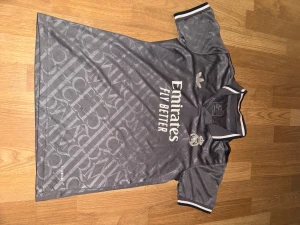 Grå Real Madrid fotbollströja Adidas (cirka XS) - Snygg grå Real Madrid fotbollströja från Adidas med vit logga och texten 'Emirates Fly Better' på bröstet. Tröjan har korta ärmar, krage med vit och mörkgrå rand samt ett diskret mönster över hela tyget. Tillverkad i lätt polyester för maximal komfort.