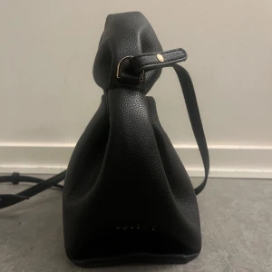 Svart handväska från Polène - Säljer en stilren svart handväska från Polène i mjukt, kornigt skinn. Väskan har en unik form med knytdetalj upptill och guldfärgade metalldetaljer. Perfekt för dig som gillar minimalistisk design och vill ha något som sticker ut. Fick i present ingen aning om den är orginal