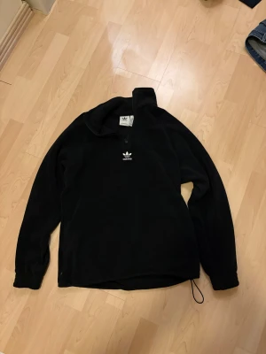 Svart quarter zip fleece-tröja från Adidas retro - Svart långärmad fleece-tröja från Adidas med halv dragkedja och broderad logga framtill. Stor Adidas-tryck på ryggen väldigt retro. Tröjan har en loose passform och snörning vid nederkanten. Perfekt för chill dagar och streetwear. Använd max 10 gånger köpt i Berlin för 80€