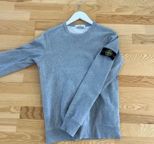 Grå Stone Island sweatshirt - Äkta Grå sweatshirt från Stone Island med klassisk rund hals och lång ärm. Ikonisk svart patch med gul och grön broderad logga på vänster ärm. Mjuk bomullskvalitet och ribbade muddar vid ärmslut och nederkant. Perfekt för dig som gillar streetwear och snygga detaljer.