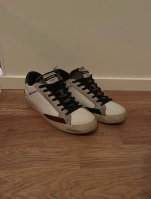 Vita sneakers från Crime London - Snygga vita sneakers från Crime London med svarta snören och detaljer i grått och vinrött. Skorna har rund tå, platt sula och är tillverkade i skinn med mockadetaljer. Märket syns tydligt på plösen och hälen. Perfekta för dig som gillar streetstyle.