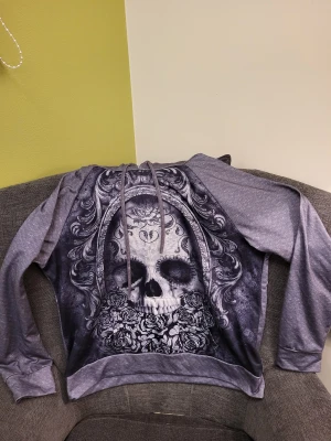Grå hoodie med döskalletryck XXL passar xl - Säljer en grå hoodie i storlek XXL med ett fett döskalletryck på framsidan, omgiven av detaljerade rosor och ornament. Tröjan har huva med snörning och långa ärmar med muddar. Materialet känns mjukt och är troligen bomull eller polyester.