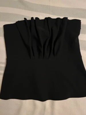  NySvart tubtopp från Gina Tricot - Säljer en svart tubtopp från Gina Tricot i storlek 38. Toppen har snygga rynkade detaljer framtill och en dold dragkedja i sidan för enkel påklädning. Perfekt att styla med jeans eller kjol för en trendig look.
