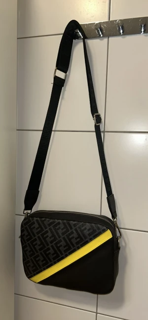 Fendi axelväska med FF-mönster och gul detalj - Snygg axelväska från Fendi i svart skinn med FF-logga i mörkgrått på framsidan och en cool gul diagonal detalj. Justerbar axelrem med Fendi-tryck och dragkedja upptill. Baksidan är helt svart och stilren. Perfekt för dig som vill sticka ut med designerstil. Kan fraktas om köparen står för kostnader 