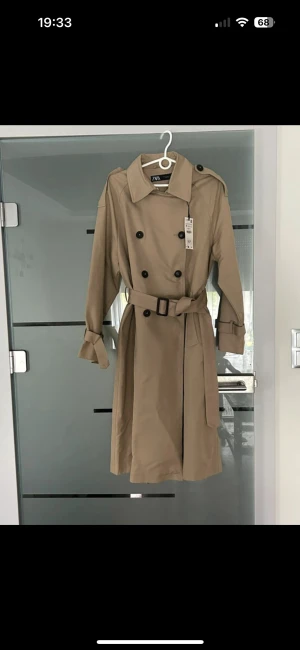 Beige trenchcoat från ZARA helt ny - Stilren beige trenchcoat från ZARA i klassisk modell med dubbelknäppning, bälte i midjan och snygga detaljer vid axlar och ärmslut. Helt ny, med etiketter, endast provat. 