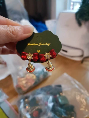 Juliga örhängen med rosett och klockor - Superfina örhängen med röda rosetter, gnistrande stjärnor och små guldiga klockor med röda detaljer. Perfekta för att maxa julstämningen och ge din look en extra festlig touch. Säljs i originalförpackning.