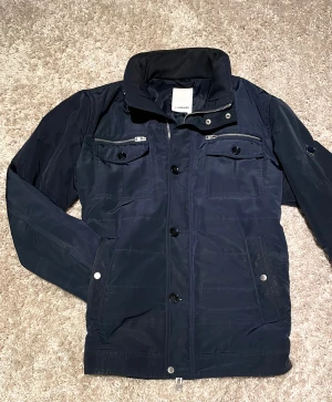 J Lindberg Field Jacket - En riktigt snygg J Lindberg Field Jacket med dun. Denna är perfekt för vinter, vår och höst. Den är mörkblå med knappast några defekter. Det går att fälla ut en luva också. 